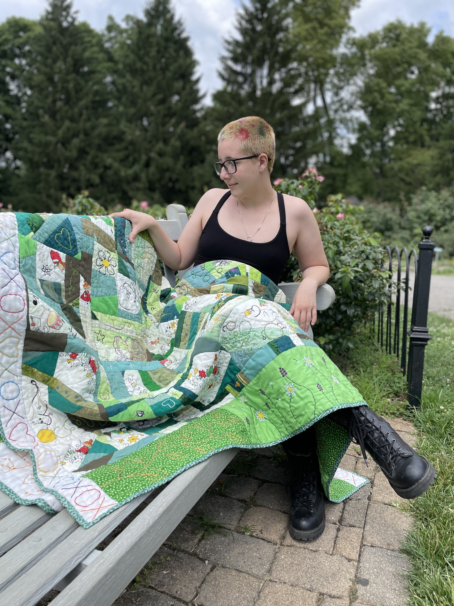 Daisy sitting in a rose garden posing with their large, patterned scrap quilt with images of Moomins, flowers, and other simple shapes. The quilt has no true pattern to it.